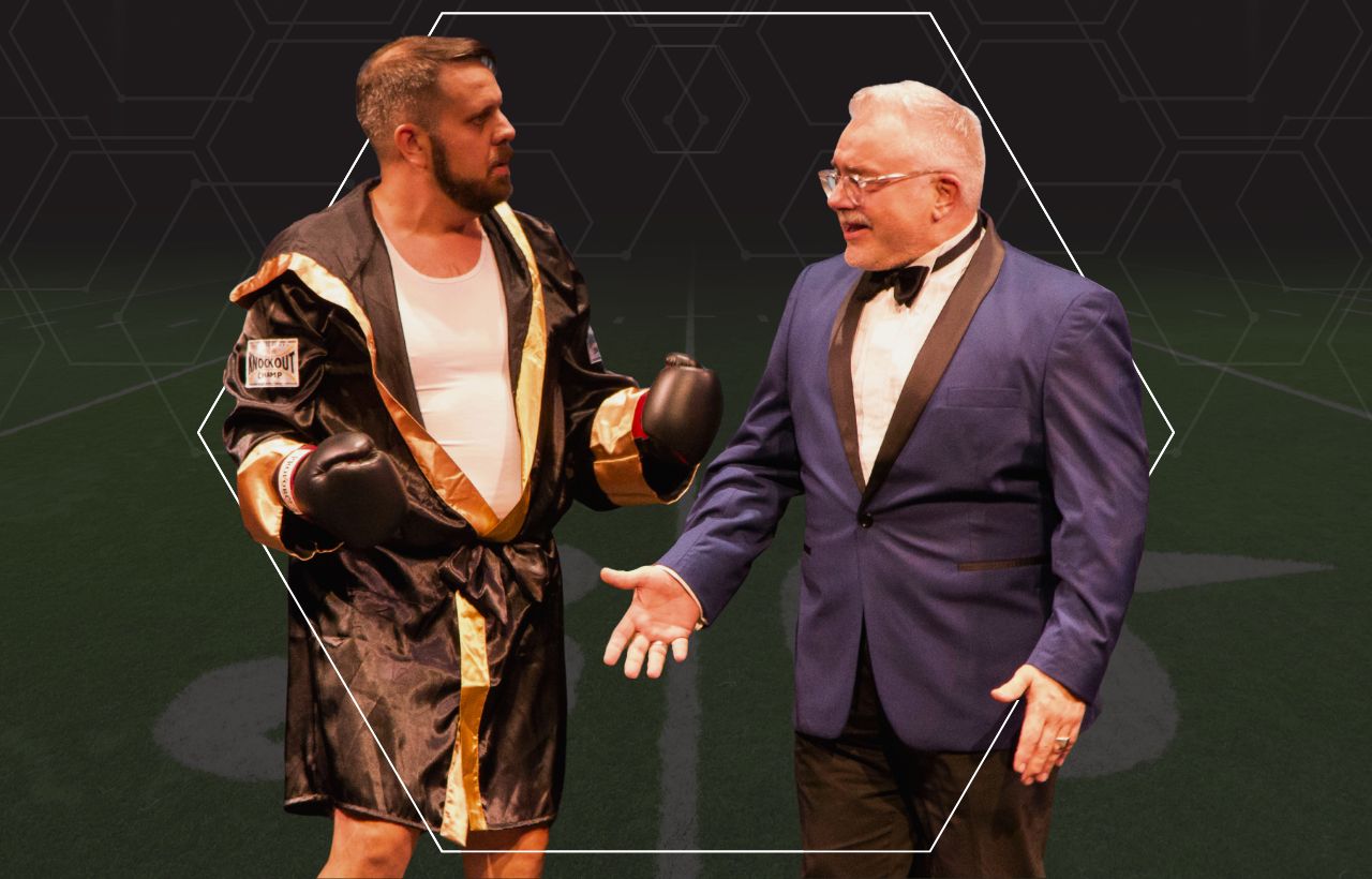 Two people stand on a football‑themed stage backdrop, one dressed in a boxing‑style robe with gold trim and the other wearing a blue tuxedo jacket.