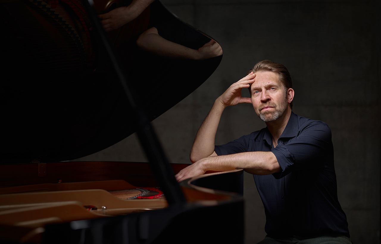 Leif Ove Andsnes seated at an open grand piano against a dark gray background, wearing a deep blue shirt, with his hand resting against his head