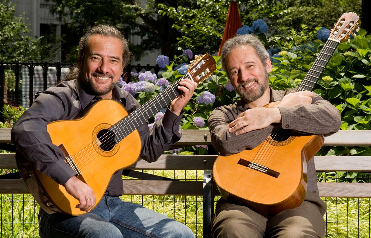 Sérgio and Odair Assad seated with their guitars on a bench outdoors