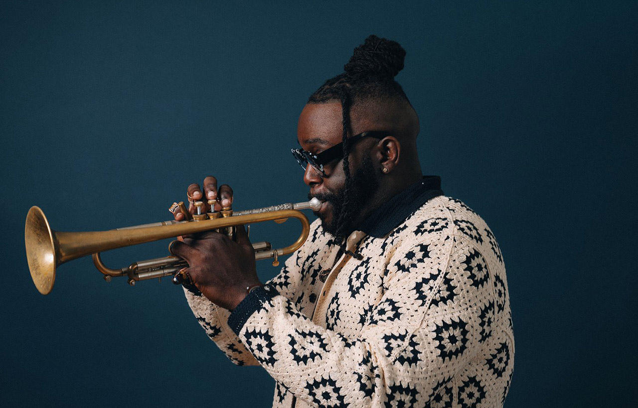 Marquis Hill wears a black and white sweater and stands playing trumpet against a blue background