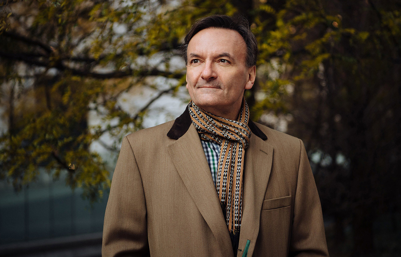 Stephen Hough stands outside in a tan coat and scarf in front of trees