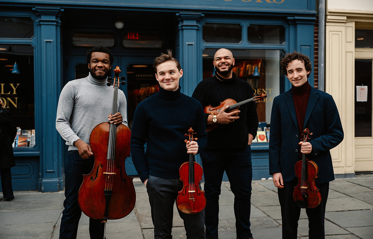 Isidore String Quartet members standing with their instruments in front of a teal building