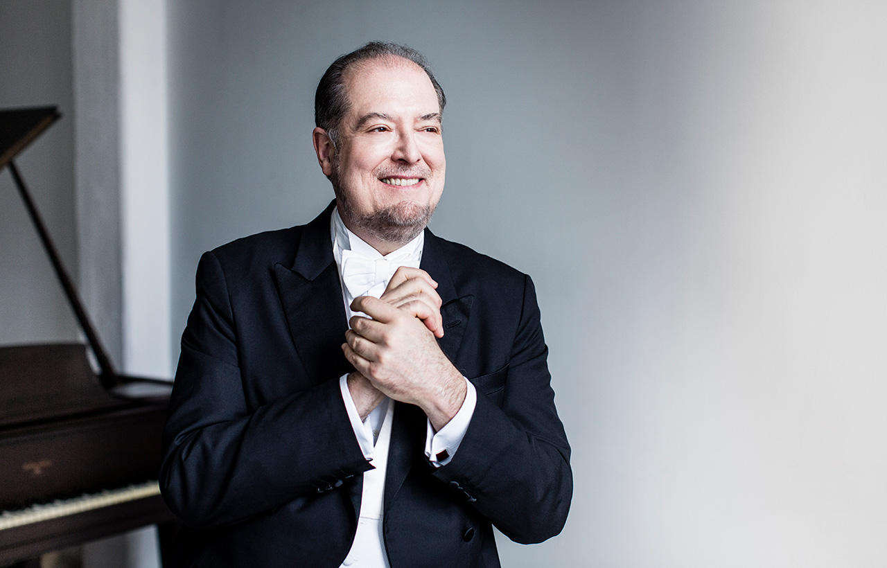 Garrick Ohlsson stands in front of a grand piano, wearing a tuxedo and clasping his hands at his chest